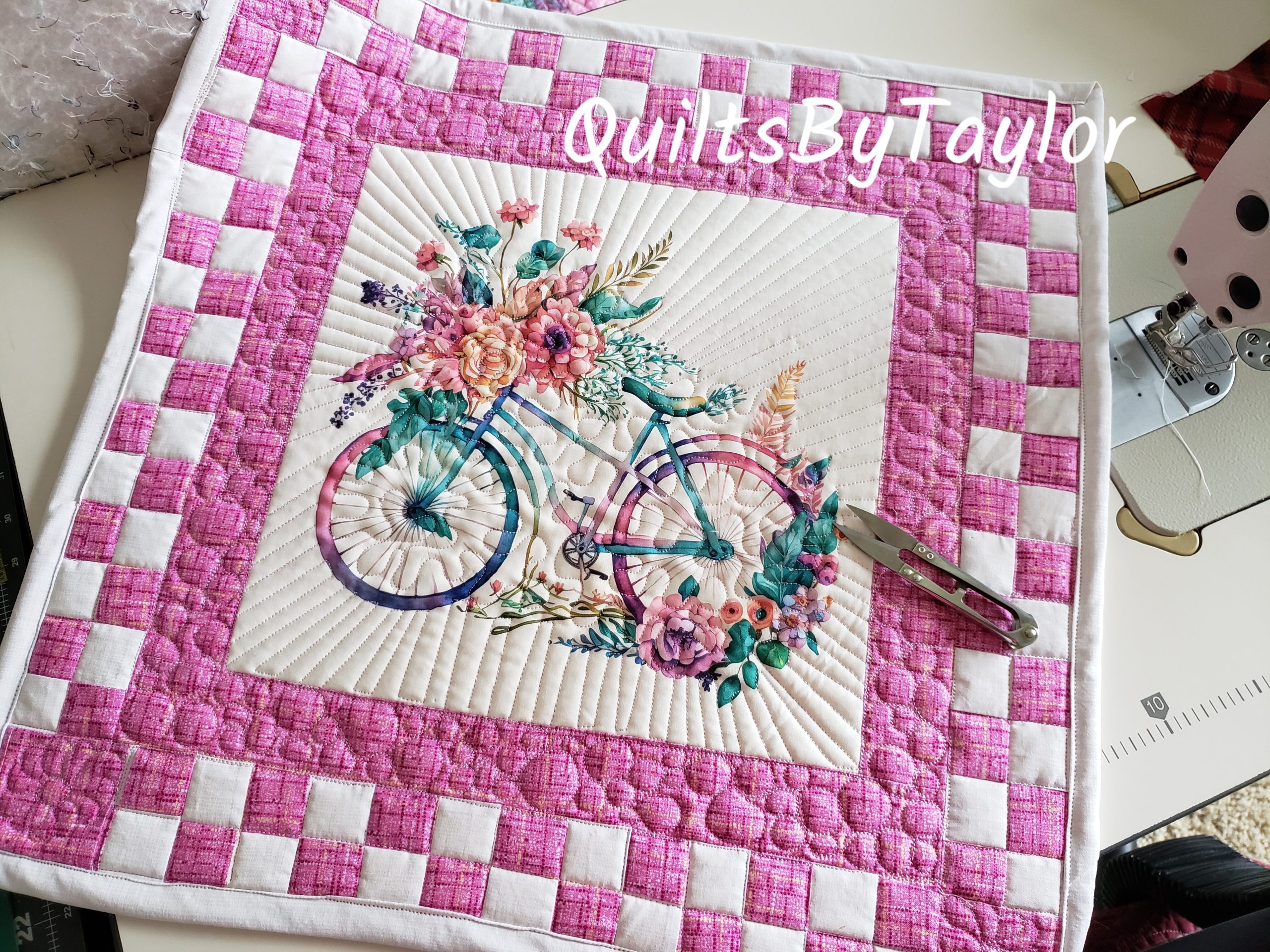 “Handmade quilted bicycle table topper with pink checkerboard border and floral bicycle center panel.